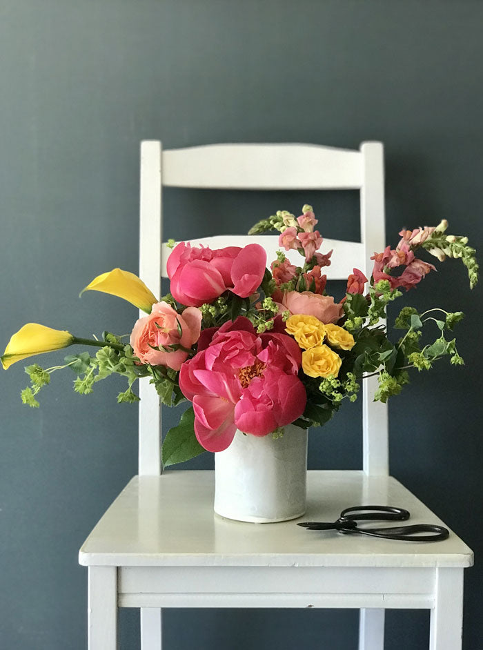 coral peonies arranged in a white pottery cylinder with peach garden roses, yellow spray roses, yellow mini callas, and orange snapdragons.  Arrangement is sitting on a white chair with a dark blue background