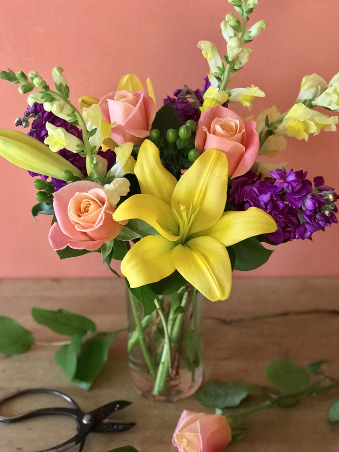 yellow lilies, purple stock, yellow snapdragons, and green hypericum berries in a clear glass vase with a peach background