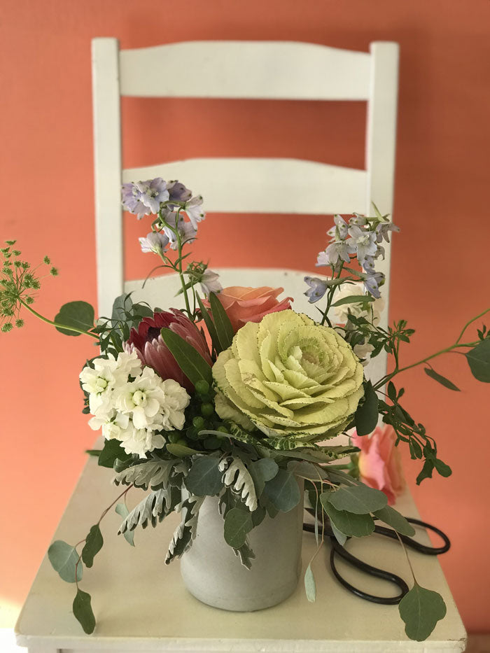 ornamental kale, pink protea, white stock, blue delphinium and peach rose in a cement vase sitting on a white chair