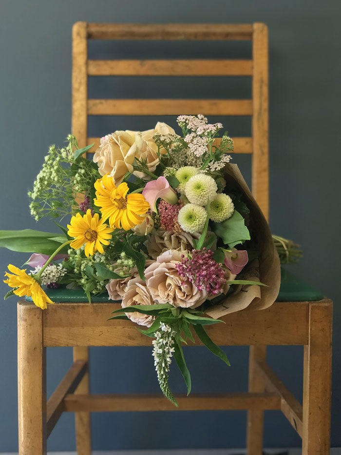 spring wrapped bouquet, locally grown flowers in yellow, peach, pink and green on a wooden chair with blue backgroun