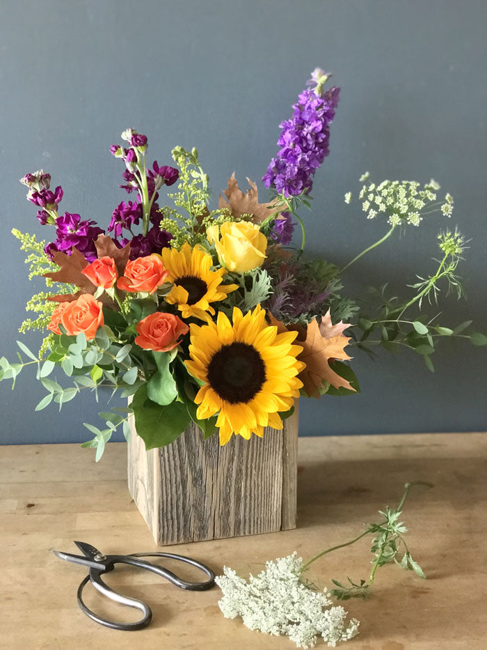 colorful fall flowers including sunflowers, larkspur, spray roses, and kale in a wooden box